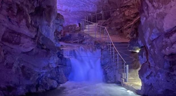 Cascata sotterranea illuminata con luce blu, scale conducono attraverso la grotta.