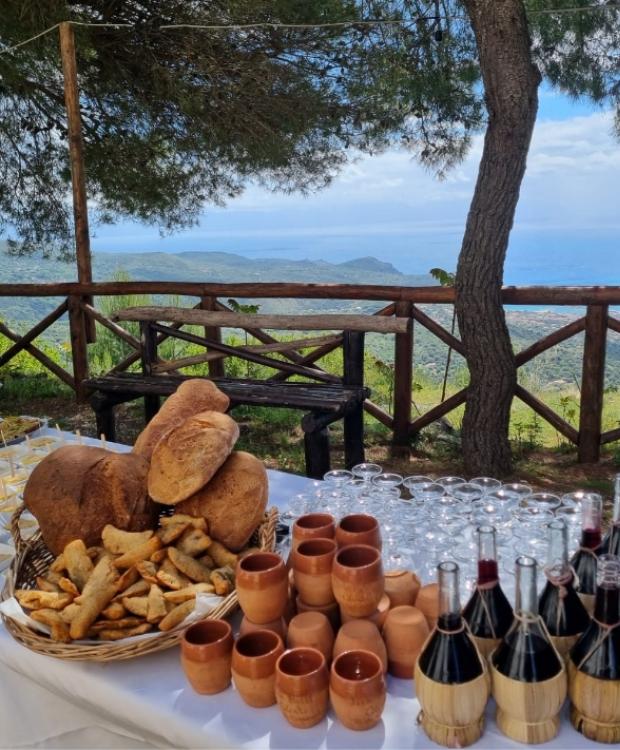 Tavola imbandita con pane, vino e vista panoramica sul mare e colline.